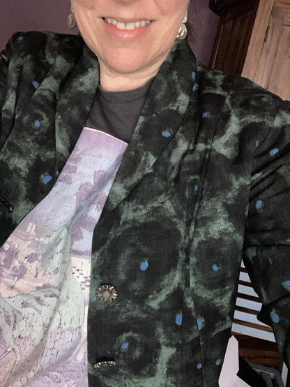 A middle aged woman wearing a tailored jacket with shawl collar in a green fabric with black and blue circles on it over a grey shop-bought T-shirt with Japanese art on it