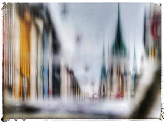 Das Bild zeigt eine Stadtansicht, die bewusst unscharf und verschwommen dargestellt ist. Links befinden sich Gebäude in verschiedenen Farben, darunter Gelb, Weiß und Blau. Rechts erkennt man eine Kirche oder ein größeres Gebäude mit grünen Türmen und einer Kuppel. Die gesamte Szene wirkt wie in Bewegung oder als künstlerische Interpretation einer Stadtansicht. Es gibt keine klaren Details, und die Farben verschmelzen ineinander, was dem Bild eine traumhafte Atmosphäre verleiht.