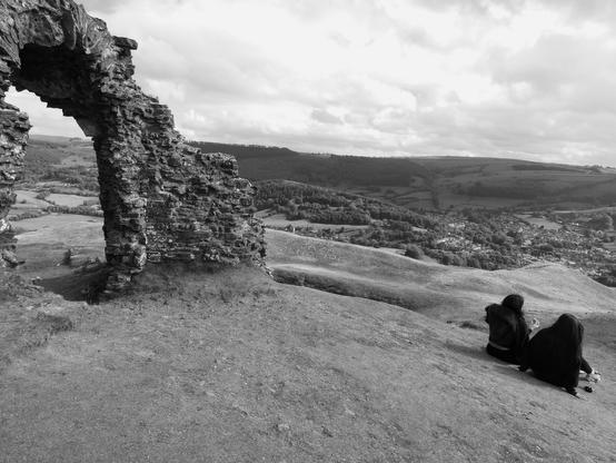 Black and white shot of part of ruined castle, a stone arch, with two young women sitting enjoying the scenery to the right, and a distant view of the message on the ramparts. Opposite side of valley and more hills, seen beyond