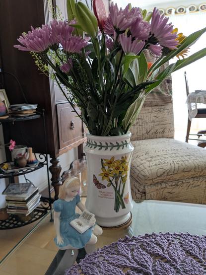 Alice - royal doulton figurine of a little girl wearing a blue dress - is reading her story under a vase with pretty purple and yellow flowers in it.
