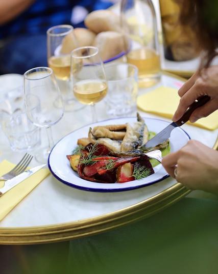 A person is cutting a plate of food with a knife and fork. The plate contains roasted beets, fried fish, and dill. There are wine glasses, bread, and butter on the table.