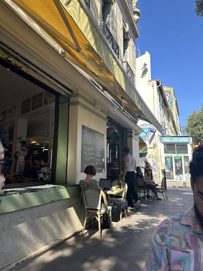 A sunny street scene with people at an outdoor cafe, one working on a laptop.