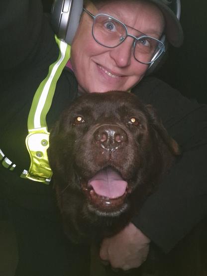 Night time selfie of a person and a chocolate brown Labrador with greying snout. The person is wearing a black jacket, yellow safety harness, red cap, silver headphones and blue-rimmed glasses. On their cap rests a headstrap torch in off position. Their free hand is around the dog's shoulders. Both are facing the camera. The dog's mouth is slightly open, showing off some blep. The person's head is resting on the dog's head. They're smiling, grateful for their time spent together.