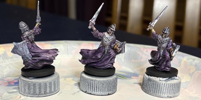 Three miniatures of skeletal warriors with a shield and a sword. They are facing in different directions and have flowing purple robes and pitted iron armor.