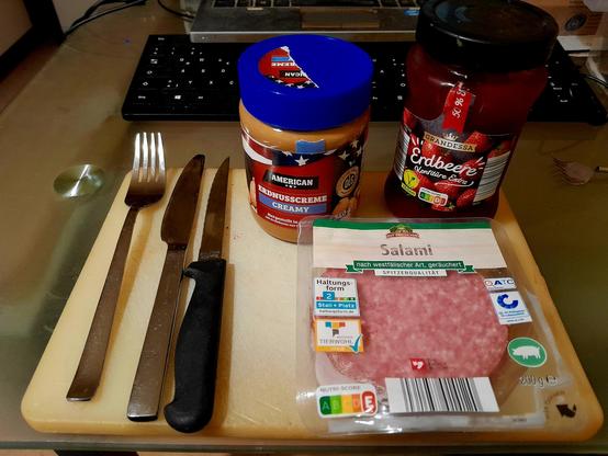 Cutlery,  peanut butter, strawberry preserves, and salami await toast toasted round side down on a cutting board.