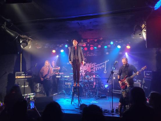 Vulture Industries performing live on a dimly lit stage. The lead singer stands dramatically on a bar stool at the center