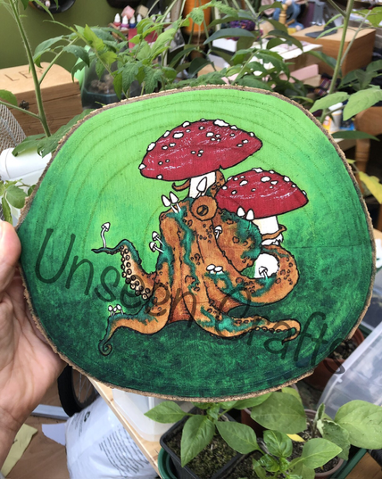 Roundish wood slice with burned and painted design showing an orange octopus sitting underneath red and white amanita mushrooms. He has tiny white mushrooms growing on him and green places that could be moss or disease, who knows 🤷‍♀️ . background painted in varying shades of green.