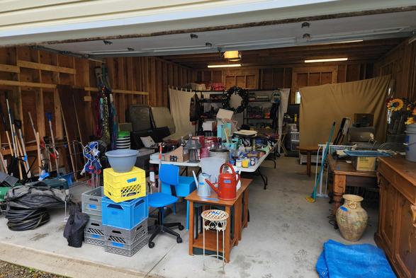 A photo of our 2 car garage full of household items that will be sold at our yard sale. There are 4 folding tables in the centre full of items and around the perimeter are shelves and furniture.