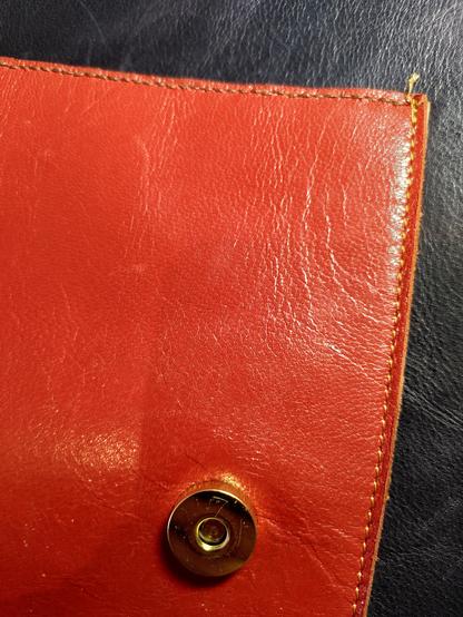 A section of red leather bag against a darker blackish coloured section of leather. There's a seam vertically on the edge of the red leather. It's stitched with a yellowish thread. Near the bottom is a round magnetic clasp. Across the top is a seam stitched with a brown thread.