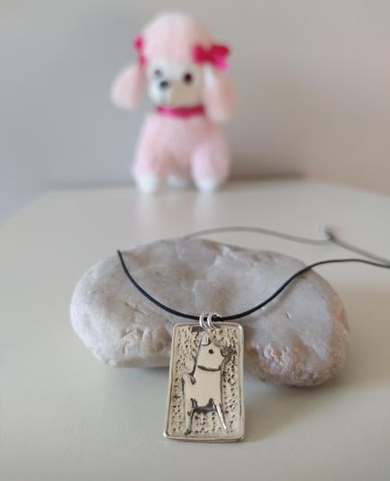 a square silver pendant with a black cord. it has the profile of a dog on it made also from silver. it's placed on a pebble and there is a puppy toy in the background.