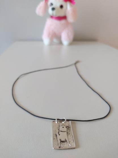 a square silver pendant with a black cord. it has the profile of a dog on it made also from silver. there is a puppy toy in the background.