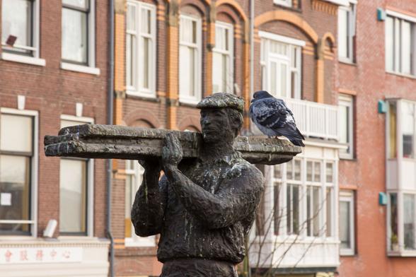 In the middle of the photo is a bronze statue of a worker with long (bronze) planks on his right shoulder. His legs are not on it. You see him from the front and from the side in work clothes with a cap. His face is friendly. At the end of the planks sits at ease, a fat, gray pigeon. The bronze worker is covered in pigeon droppings. In the background, more vague, rows of The Hague houses of brown-red bricks with many windows, with white window frames. You see the pigeon from behind, but his head is turned a little and looks backwards with a crooked eye. As if he is annoyed that I am photographing it.