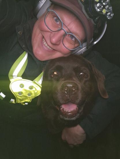 Night time selfie of a person and a chocolate brown Labrador with greying snout. The person is wearing a black jacket, yellow safety harness, red cap, silver headphones and blue-rimmed glasses. On their cap rests a headstrap torch in off position. Their free hand is around the dog's shoulders. Both are facing the camera. The dog's mouth is slightly open, showing off some blep. The person's head is resting on the dog's head. They're smiling, grateful for their time spent together.
