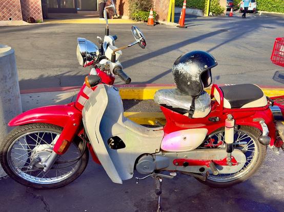 Here is a photo of a red and white motor scooter with a beaten up helmet tied to the seat parked in a TJs parking lot. There is so much duct tape in important areas of the bike, it appears to be constructed using the material. It is as amazing as it is scary.