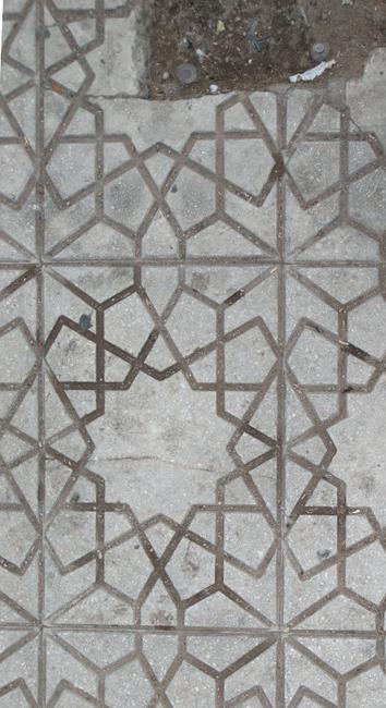 A top-down photograph of grey rectangular street paving. Each paver has a 12-pointed star in the centre surrounded by kites and different kinds of pentagons. 
The pavers are grimy and worn from use over time. Two of the pavers are broken: brown earth and metal pins are visible.