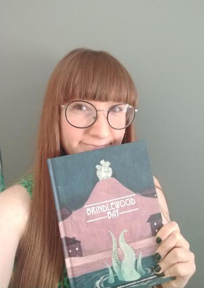 Ich, blass, rotbraune Ponyfrisur und große runde Brille, halte ein Buch, das Regelwerk von Brindlewood Bay, in die Kamera.