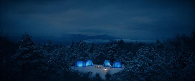 A clearing in the snow-covered forest contains four tents that are illuminated from the inside. Tall, triangular, and blue, they surround two fires. It looks very cozy.