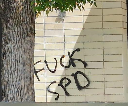 "Fuck SPD" spraypainted in black on the side of a tan brick wall, next to a tree.