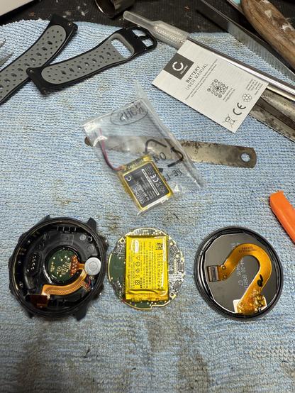 An open electronic device, likely a smartwatch, is displayed on a blue cloth. The internal components are exposed, showcasing circuit boards and a battery. Nearby items include straps, a user manual, a plastic bag containing a new battery, and various tools.