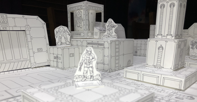 A photo showing a papercraft sci-fi scene.  Blocky, white forms create a space station interior with black line art on the walls and floors. Some papercraft miniature standees populate the scene resembling space-suit-wearing soldiers.