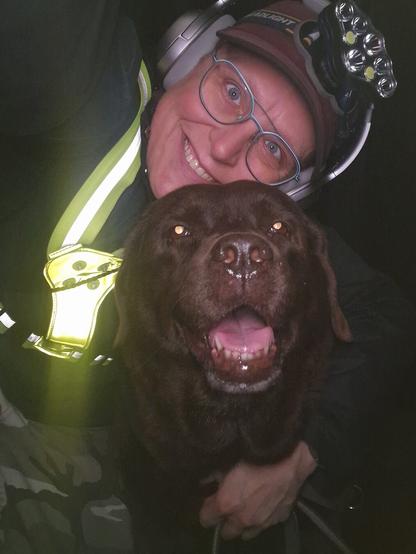 Night time selfie of a person and a chocolate brown Labrador with greying snout. The person is wearing a black jacket, yellow safety harness, red cap, silver headphones and blue-rimmed glasses. On their cap rests a headstrap torch in off position. Their free hand is around the dog's shoulders. Both are facing the camera. The dog's mouth is slightly open, showing off some blep. The person's head is resting on the dog's head. They're smiling, grateful for their time spent together.