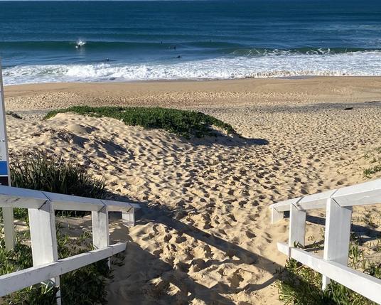 White wooden railings on either side of the beach access (can't see the steps, just the sand). The light coloured sand is dimpled with footsteps. The ocean is glassy blue with a tinge of green. A surfer is just starting down the face of a wave, still belly down. Two surfers are paddling out to where others wait for the next wave.