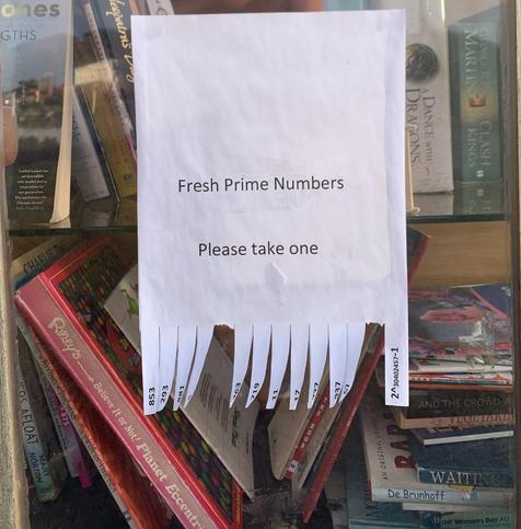 A piece of plain white paper is stuck to a glass door, behind which are a mess of books on shelves. Printed on the paper in a sans serif font is "Fresh Prime Numbers. Please take one". The bottom edge of the paper has been cut into strips, with a prime number written in each. Two have been taken.