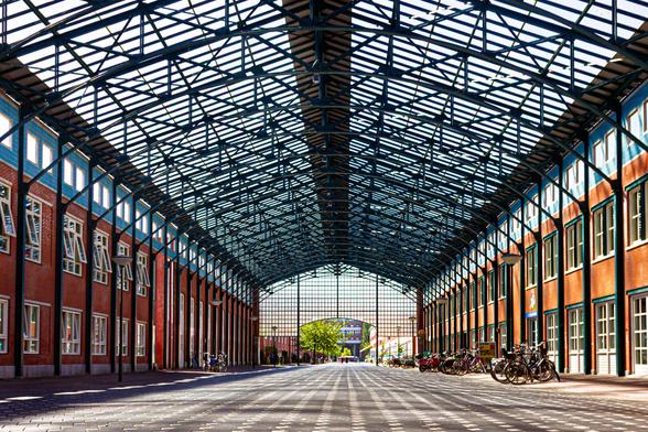 A large hall, with a roof of steel trusses, which deliberately takes up a large part of the top of the photo. On the left and right are colorful facades of houses (pastel blue and green with orange bricks). In the distance, through the opening on the other side, you can see the green of trees and more houses. The wide floor is covered with light gray paving stones, and through them horizontal, white triangular lines of white bricks. The light from the roof falls on it very nicely.