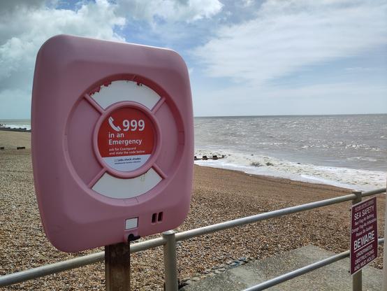 A red sun-faded lifebuoy by railings next to a shingle beach. Grey sea and a partly cloudy sky.