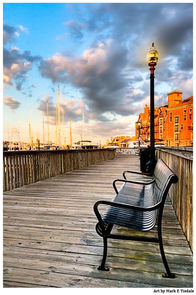 Photo of a wooden boardwalk along Boston’s Harborwalk at sunset, with a single empty bench, a lamppost, warm light on nearby brick buildings, and a marina view.
