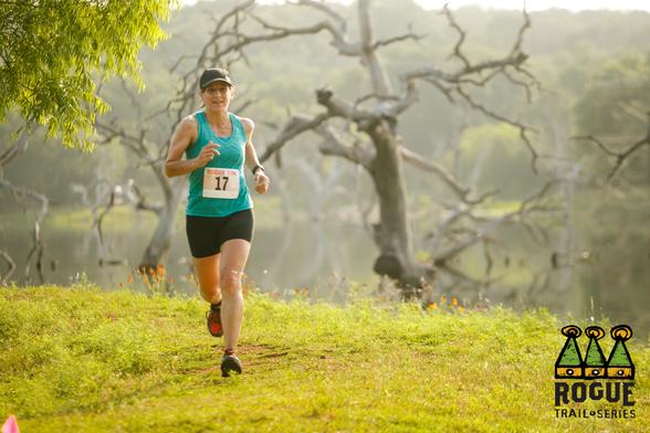A person is running on a grassy trail near a body of water, wearing a turquoise tank top, black shorts, and a black cap. They have a race bib number 17 pinned to their top. In the background, there are leafless trees and reflections on the water. The foreground has a few scattered flowers. There is a logo in the bottom right corner that reads "ROGUE TRAIL SERIES."