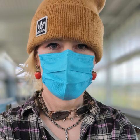 A photo of Alice at a train terminal, wearing a caramel-colored beanie and pink/black/white plaid shirt. They're wearing a sky blue mask and strawberry earrings, with two steel chain necklaces visible below the sunglasses clinging to their neck. Blonde hair juts out from beneath the beanie, framing one side of Alice's face and their dark blue eyes.