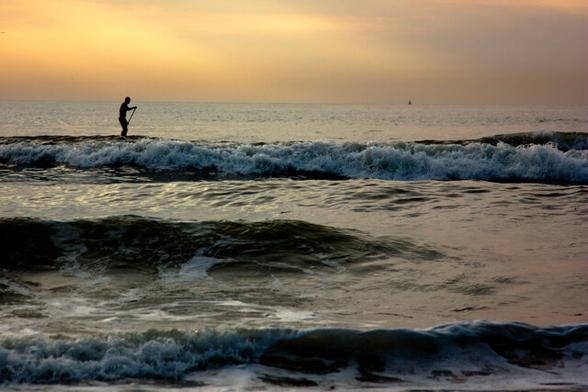 A surfer at sea during the golden hour. Two thirds of the photo is sea, above that an orange sky. A little below the horizon a long foaming wave horizontally across the photo. With on the left of it a lonely surfer, standing and paddling on a surfboard. In the foreground halfway a wave that is coming in and at the very bottom over the entire length, horizontally, a low, foaming wave.