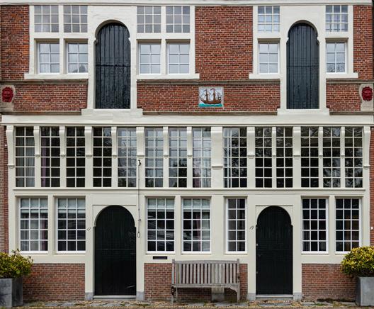 The facade of a beautiful old building in Hoorn. Two floors are photographed from the front. There are four identical, green, wooden doors with a round top. Two on the first floor and two on the ground floor. The ground floor has windows from one meter from the ground to the second floor over the entire length. They are two rows of narrow, rectangular windows with white styles divided into small panes. The two doors are between them. On the second floor, next to the two doors, there are the same narrow windows with small panes but fewer. Four on both sides of the left door and two on each side of the right upper door. In the middle above the upper doors a tile with a colored sailing ship. On the right and left at the top of the facade two red painted lion heads.
