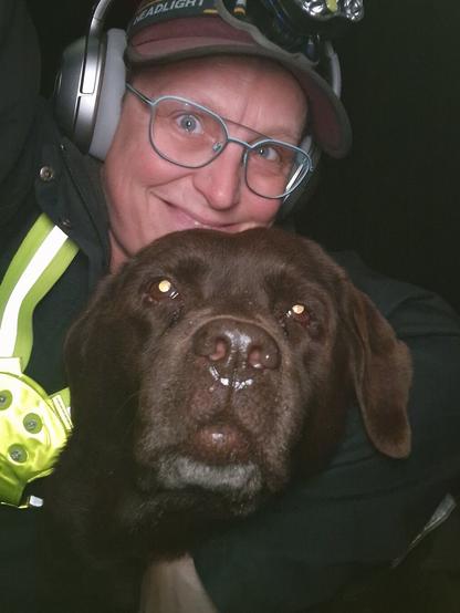 Night time selfie of a person and a chocolate brown Labrador with greying snout. The person is wearing a black jacket, yellow safety harness, red cap, silver headphones and blue-rimmed glasses. On their cap rests a headstrap torch in off position. Their free hand is around the dog's shoulders. Both are facing the camera. The person's head is resting on the dog's head. They're smiling, grateful for their time spent together.