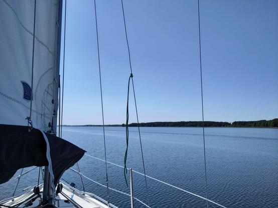 Bow of a sailing yacht with sails up, sailing across a relatively calm lake. Shore with trees visible in the distance.
