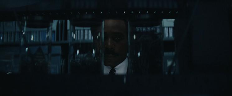 Inside a large storage room, Mr. Milchick’s face is partially obscured by tall, clear containers containing paper clips. He stares at one with muted hostility on his carefully composed  face.