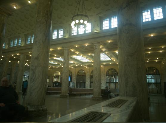 The passenger waiting hall of the Utica Amtrak station.  Marble pillars hold up an arched skylight, presiding over rows of benches. At the far end, you can see the "now serving" display of the Utica DMV