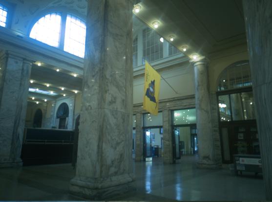 The platform passageway, presided over by several rectangular marble columns. A large yellow flag flies over the glass doors leading to the tracks.