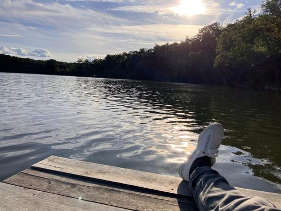 A close-up view of a person’s leg and white sneaker resting on the edge of a wooden dock. The serene lake reflects the sky and sunlight, with trees lining the opposite shore.