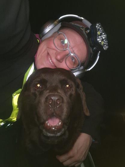 Night time selfie of a person and a chocolate brown Labrador with greying snout. The person is wearing a black jacket, yellow safety harness, red cap, silver headphones and blue-rimmed glasses. On their cap rests a headstrap torch in off position. Their free hand is around the dog's shoulders. Both are facing the camera. The dog's mouth is slightly open, showing off some blep. The person's head is resting on the dog's head. They're smiling, grateful for their time spent together.