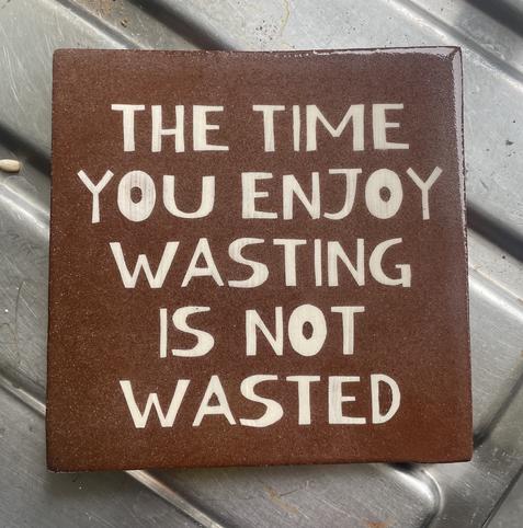A handmade terracotta clay tile decorated using a hand cut stencil and sgraffito, with the words ‘The time you enjoy wasting is not wasted’.