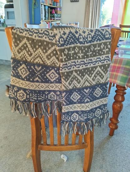 A scarf draped over the back of a chair: it is knitted with navy, light grey, and dark grey in a series of intricate colourwork patterns, and fringed at each end.