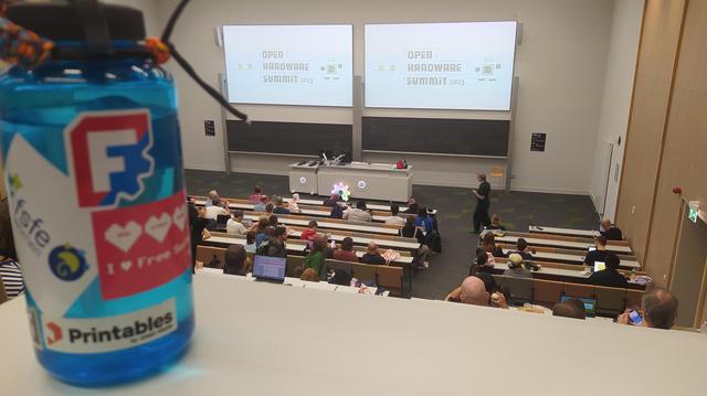 A lecture hall, the OHS branding on the screens. My waterbottle is photobombing with freecad stickers and fsfe stickers..