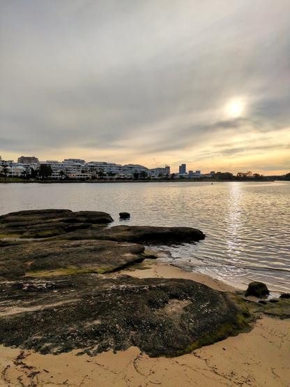 The water of Sydney Harbour washing up on the sand and rocks. Apartments on the far shore are visible in the distance. The golden sky on a cloudy day is reflected in the water.