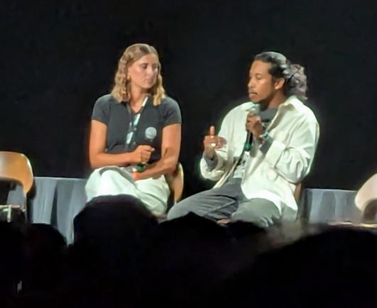 A white woman with shoulder length blond hair on the left. A black man on the right with his hair in a ponytail. They're on a stage talking