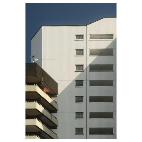 Ein weißes Wohn-Hochhaus. Fenster und stapeln sich wie Kisten übereinander. Versetzt davor ein weiteres Hochhaus aus Waschbeton. Beige Balkonbrüstungen umlaufen das Gebäude. Eine einsame rote Satellitenschüssel durchbricht die monotonen Linien.
