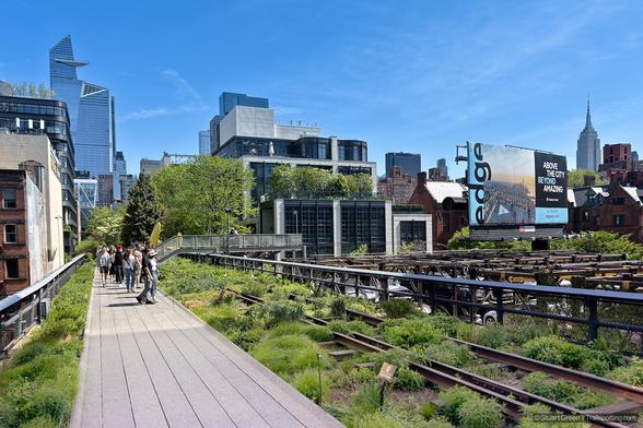 An elevated view of the High Line in New York City, showcasing its unique blend of urban architecture and greenery. A walkway lined with plants stretches through the scene, with pedestrians strolling along the path. In the background, modern skyscrapers dominate the skyline, including Hudson Yards' distinctive towers. Tthe Empire State Building stands tall in the distance, grounding the image in NYC’s iconic landscape.