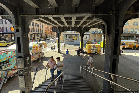 The end of the High Line in New York City, captured from the top of a staircase leading down to street level. The elevated walkway features steel beams and rivets, reflecting its industrial past as a former rail line. Below, food trucks line the sidewalk, serving a variety of meals and drinks to pedestrians. People are seen walking up and down the stairs, while others gather around the vendors, contributing to the lively, bustling atmosphere.