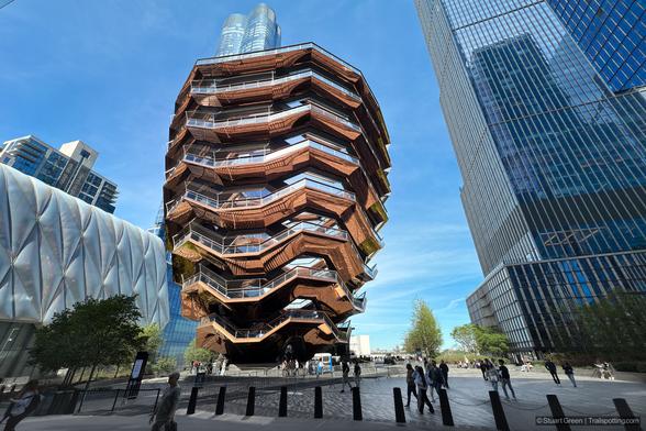 The Vessel, a striking architectural centerpiece in Hudson Yards, New York City, stands prominently in this image. Its honeycomb-like structure consists of interconnected staircases and platforms, forming a geometric labyrinth of reflective copper-colored surfaces. Surrounding the Vessel are sleek modern skyscrapers, including the Shed, a cultural center distinguished by its expandable shell. The urban environment is vibrant, showcasing the fusion of innovative architecture and contemporary city life.